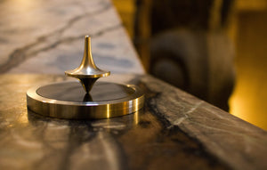 A top-quality, lustrous brass spinning top perfectly balanced on a mirror-finish metal base, epitomizing the pinnacle of craftsmanship. This image symbolizes the exceptional standard of products offered by KME, the premier online destination for the discerning shopper with visual impairments or those with low-bandwidth connections.